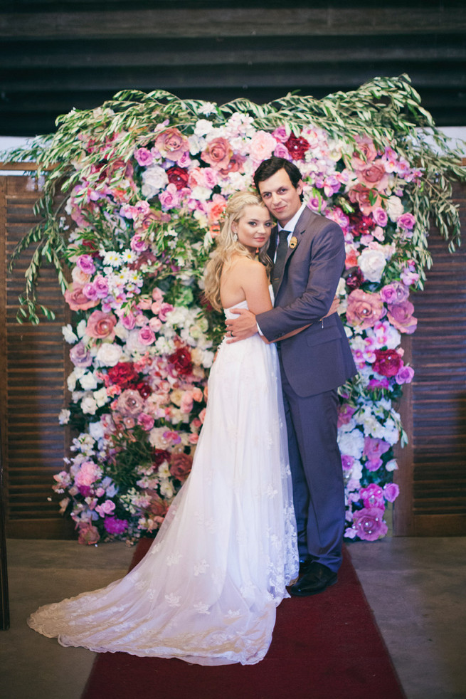 Stunning floral backdrop in pink and red. Groom in grey suit with grey tie. Green White Rustic South African Wedding // Justin Davis Photography